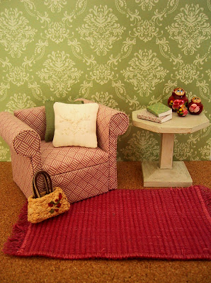 Modern dolls' house miniature scene with green and cream patterned walls, a pink and white armchair and white side table on a pink woven floor rug.