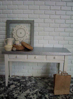 Modern dolls' house white and zinc side table, displayed against a white brick wall, with a selection of white, grey and natural miniature accessories displayed on top.
