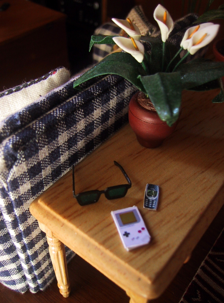Modern dolls' house miniature side table behind a blue and white checked sofa. On the table is a potted lilly plant, a pair of sunglasses, a Gameboy and a Nokia mobile phone.