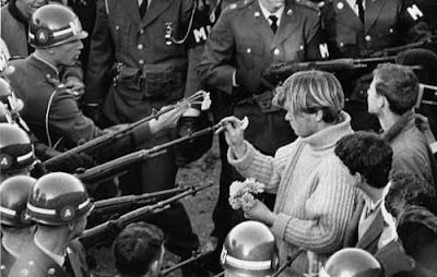 1967 black and white photo of a young blond man placing a flower into the end of a National Guard soldier's rifle