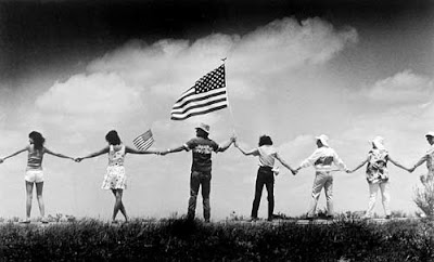 Black and white photo of people standing in a rural area, holding hands, America flag near the center, shot from a distance behind them