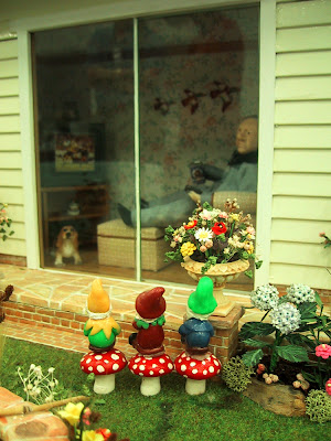 One-twelfth scale miniature scene of a man inside a house with a beer in his hand watching a football match on TV. Outside the ranch sliders in the garden, three gnomes are lined up sitting on mushrooms, watching the match through the glass.